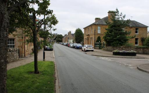 A central road lined with cars. Light brown buildings line either side of the road.
