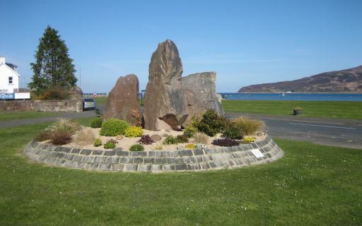 A monument consisting of three large stones atop a bed of beige coloured gravel and green shrubs. The Holy Isle can be seen across a thin body of water in the top right quarter of the image