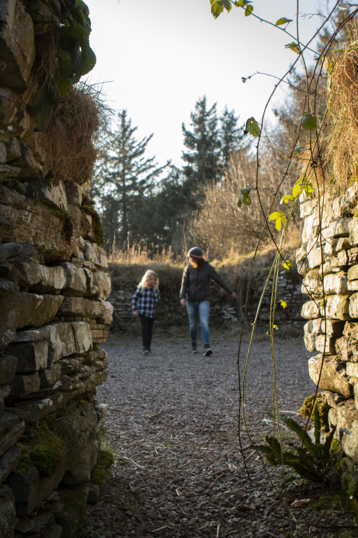 Young girl and mother walk between two thick stone walls with overhanging vines.