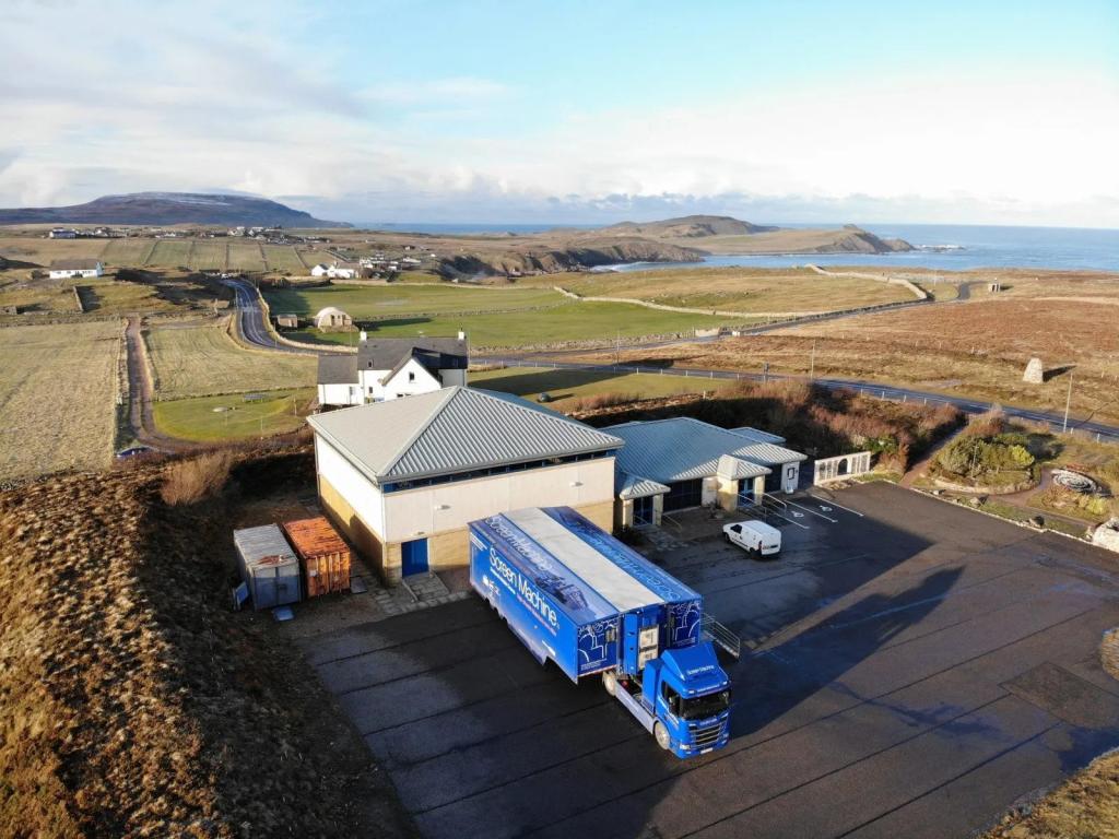 A large blue articulated lorry, with white font which reads 'Screen Machine' on its side, is parked on tarmac outside of a white and light brown building with a grey roof. Rolling green fields and white houses can be seen in the distance.