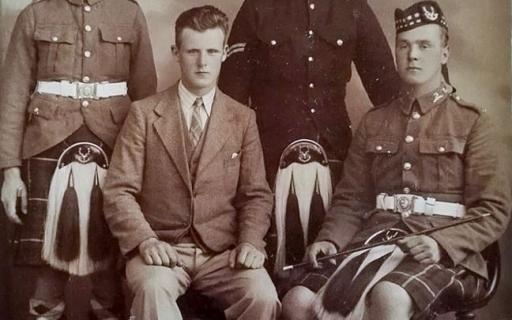 Three men in army uniforms and kilts along with a man in a suit, seated, pose for a photograph.