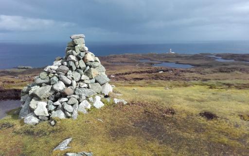 A large grey stone cairn is stacked on top of a mountain, looking out towards the sea on a cloudy day.