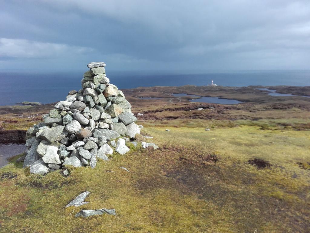 A large grey stone cairn is stacked on top of a mountain, looking out towards the sea on a cloudy day.