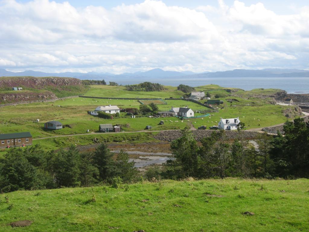 Looking onto grassy fields with scattered white and brown buildings in the distance. Green leaved trees line the foreground.