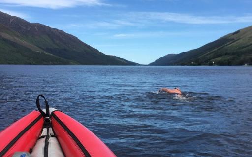 A swimmer swims in the waters of a loch. The front end of a red canoe boat can be seen in the bottom left of the image. In the distance, hills can be seen flanking the loch.