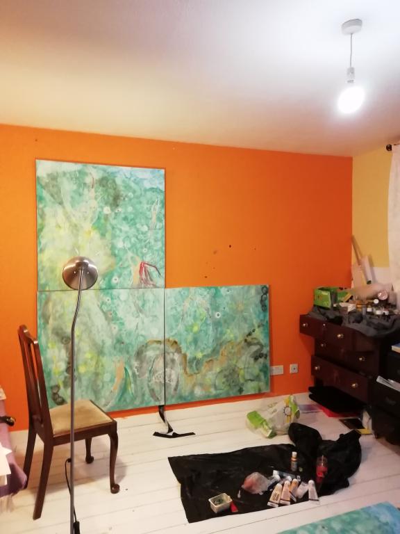 Three painted canvases against an orange wall. The three canvases are painted in shades of blue, green, and white to represent a seascape. A brown chair and art supplies sit in front of the canvases.