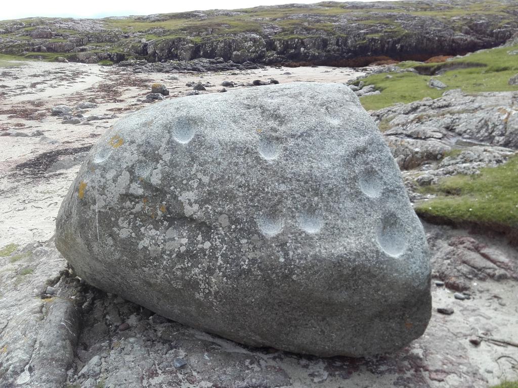 A large grey stone stands in the centre of the image. Large cup-marks are visible on the surface of the stone.