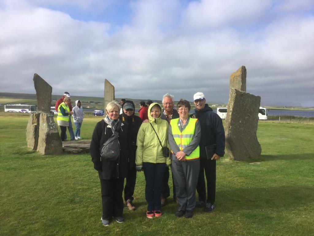 A group of five tourists stand at the centre of the image with a tour guide who is wearing a high vis vest. The group are standing in front of the Standing Stones of Stenness.