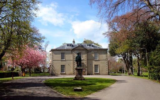 The front facade of Nairn Museum with a large statue of Dr. Grigor on plinth. Large trees and a cherry blossom tree with pink flowers line the driveway up to the museum.