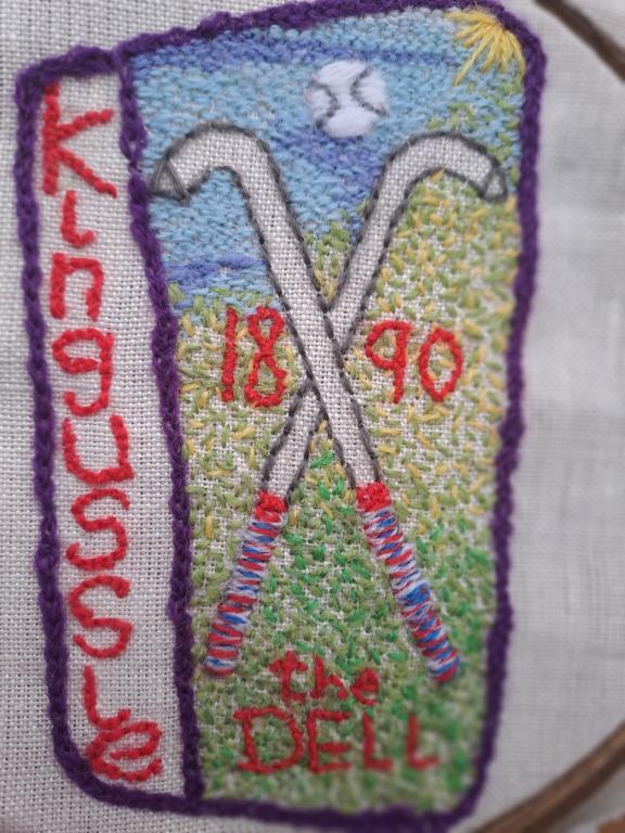 Two shinty camans are crossed in the centre of the stitched piece. The words 'Kingussie', 'the DELL' and the year '1890' are stitched in red.