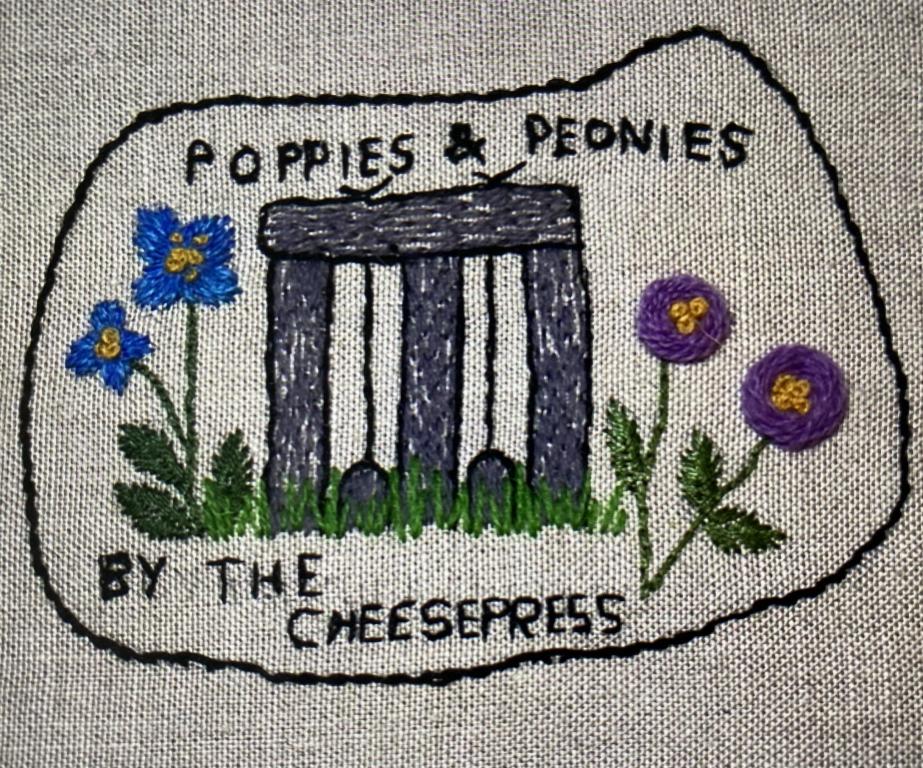 Blue and purple flowers stitched either side of a grey cheese press. The words 'Poppies and Peonies by the Cheesepress' are stitched in black above and below the cheese press.