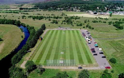 An aerial view of a large, green shinty pitch. Cars line the concrete on the right side of the pitch. A river curves around a bank to the left hand side of the pitch.