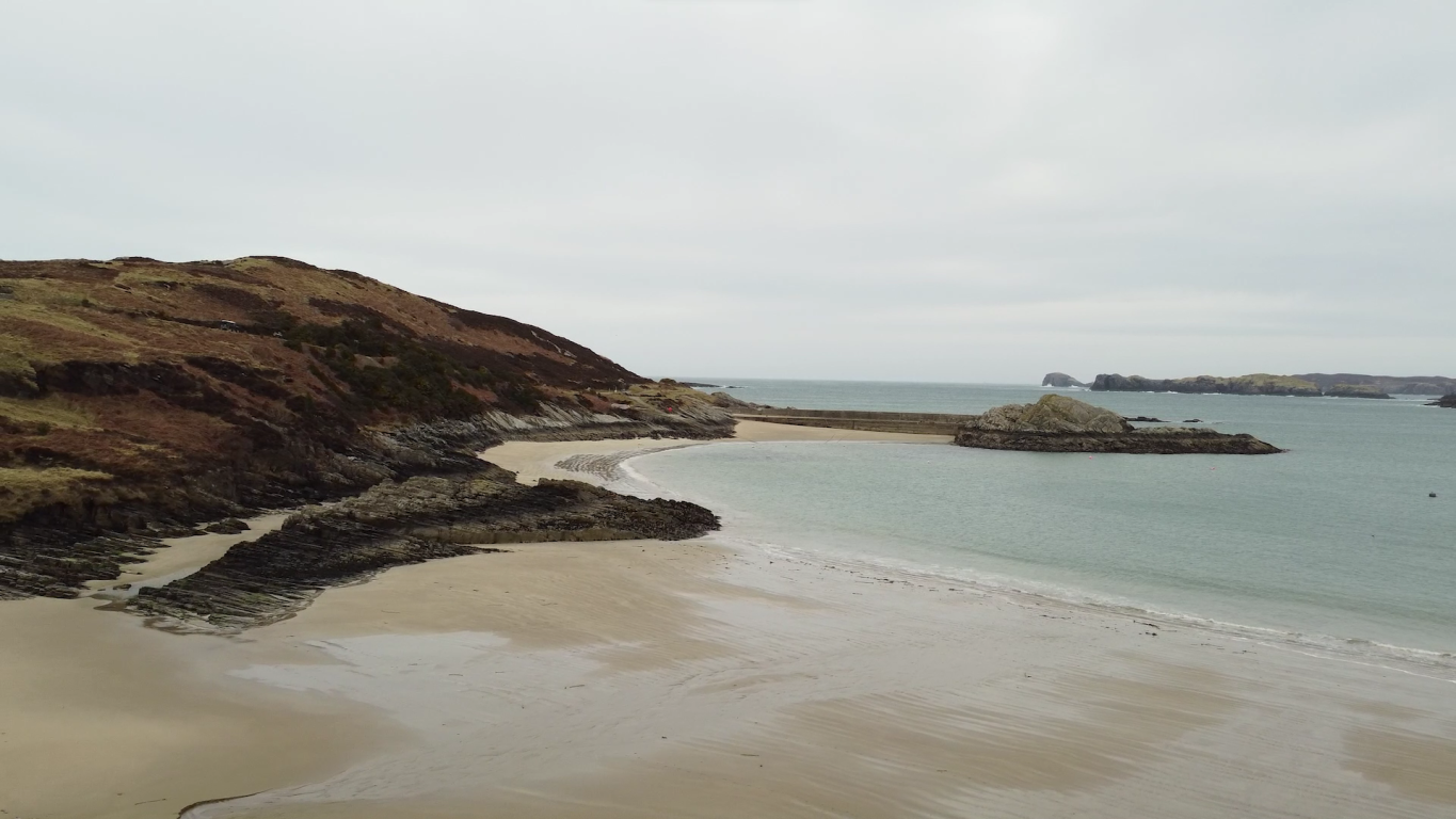 Pale yellow sands of a beach meet the light blue waters of the sea on the right hand side of the image. In the background, a hill slopes down to the shoreline. A causeway leading to an island can also be seen in the distance.