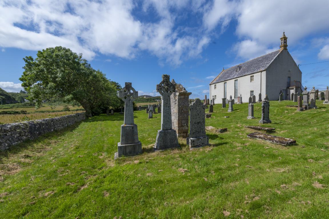 In the foreground of the image gravestones, including two Celtic crosses, are visible. A large white building with a grey roof can be seen in the background, on the right hand side of the image. A low stone wall runs parallel to a line of trees on the left hand side of the image.