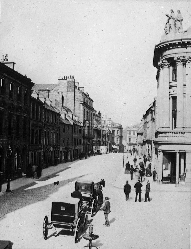 Inverness High Street in 1870. A small dog is seen in the centre of the High Street, along with a horse and carriage. People walk down the pavement on the right hand side of the image. Inverness High Street in 1870. A small dog is seen in the centre of the High Street, along with a horse and carriage. People walk down the pavement on the right hand side of the image.