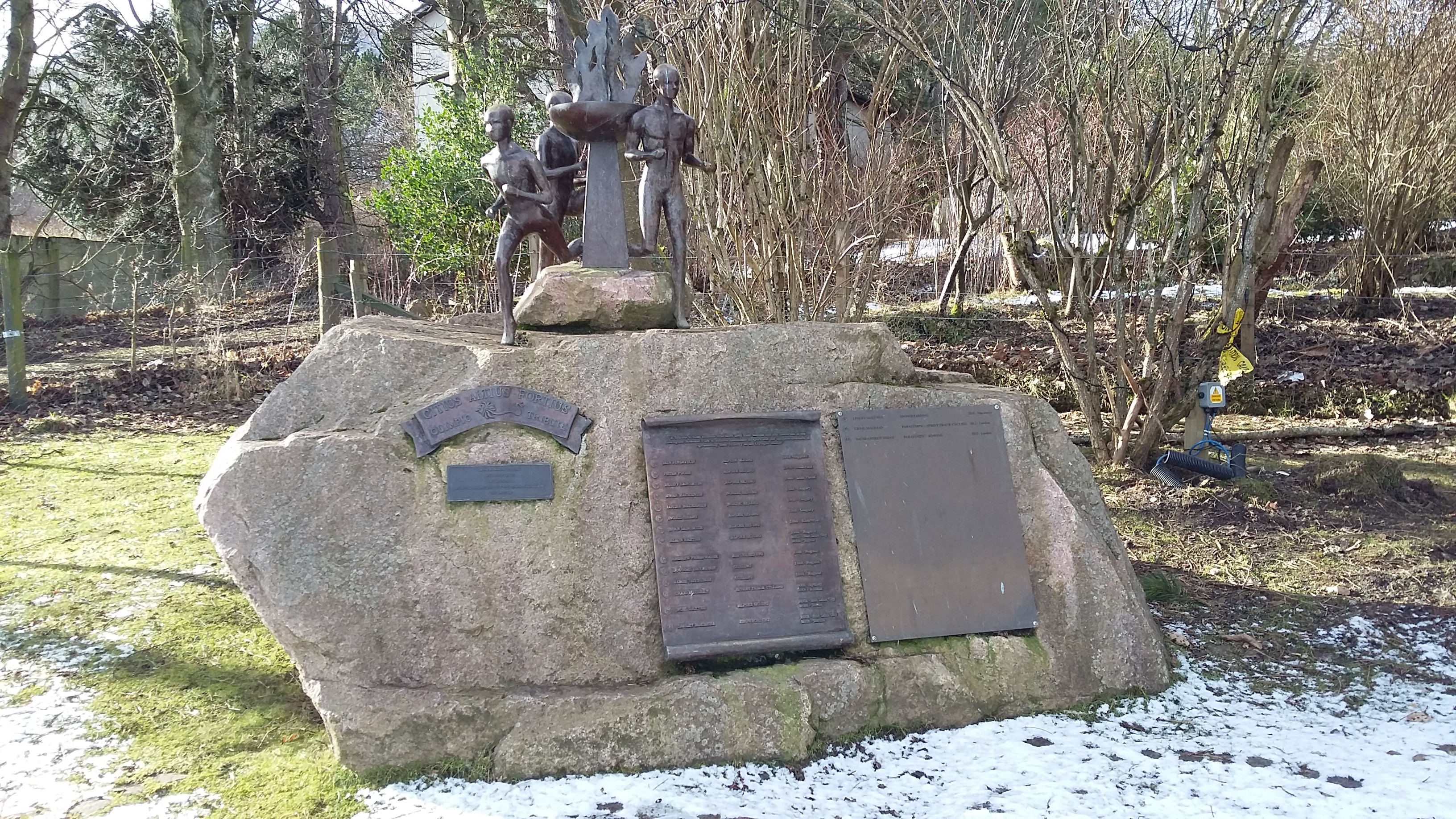 A monument with three figures surrounding the Olympic flame, cast in bronze, stand atop a carved stone block. Two plaques, also cast in bronze, with the names of olympians from the area are attached to the memorial sculpture