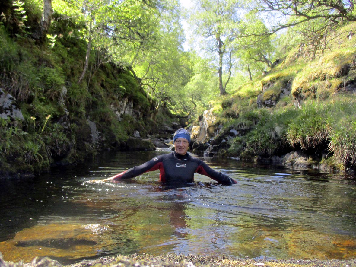The submitter in a blue swim cap and red and black wetsuit, Stephen Austin, swimming in the clear water of a gorge surrounded by trees