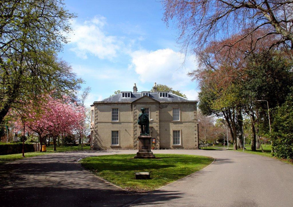 The front facade of Nairn Museum with a large statue of Dr. Grigor on plinth. Large trees and a cherry blossom tree with pink flowers line the driveway up to the museum.